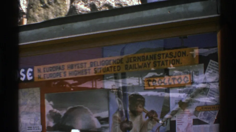 1976 Norway 8mm frame showing a bilingual railway-station sign through glass, with reflections and a chimpanzee poster beneath it.