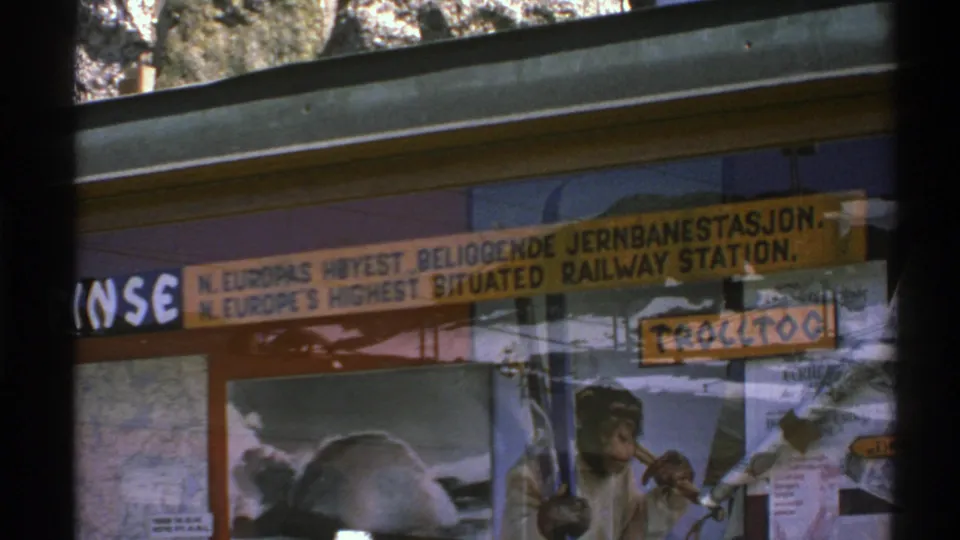 1976 Norway 8mm frame showing the yellow highest-situated railway-station sign through reflective glass above poster material.