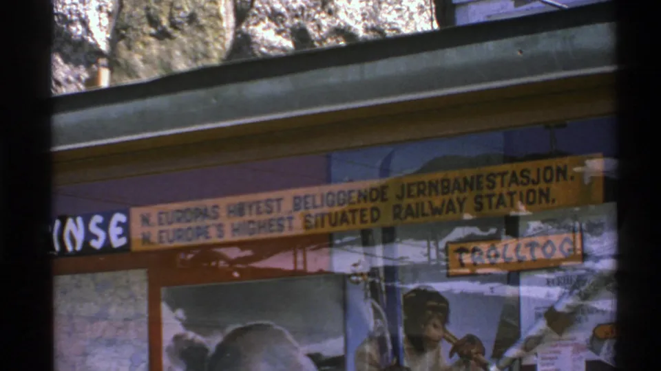 1976 Norway 8mm frame capturing an angled train-window view of the bilingual station sign and chimpanzee poster display.