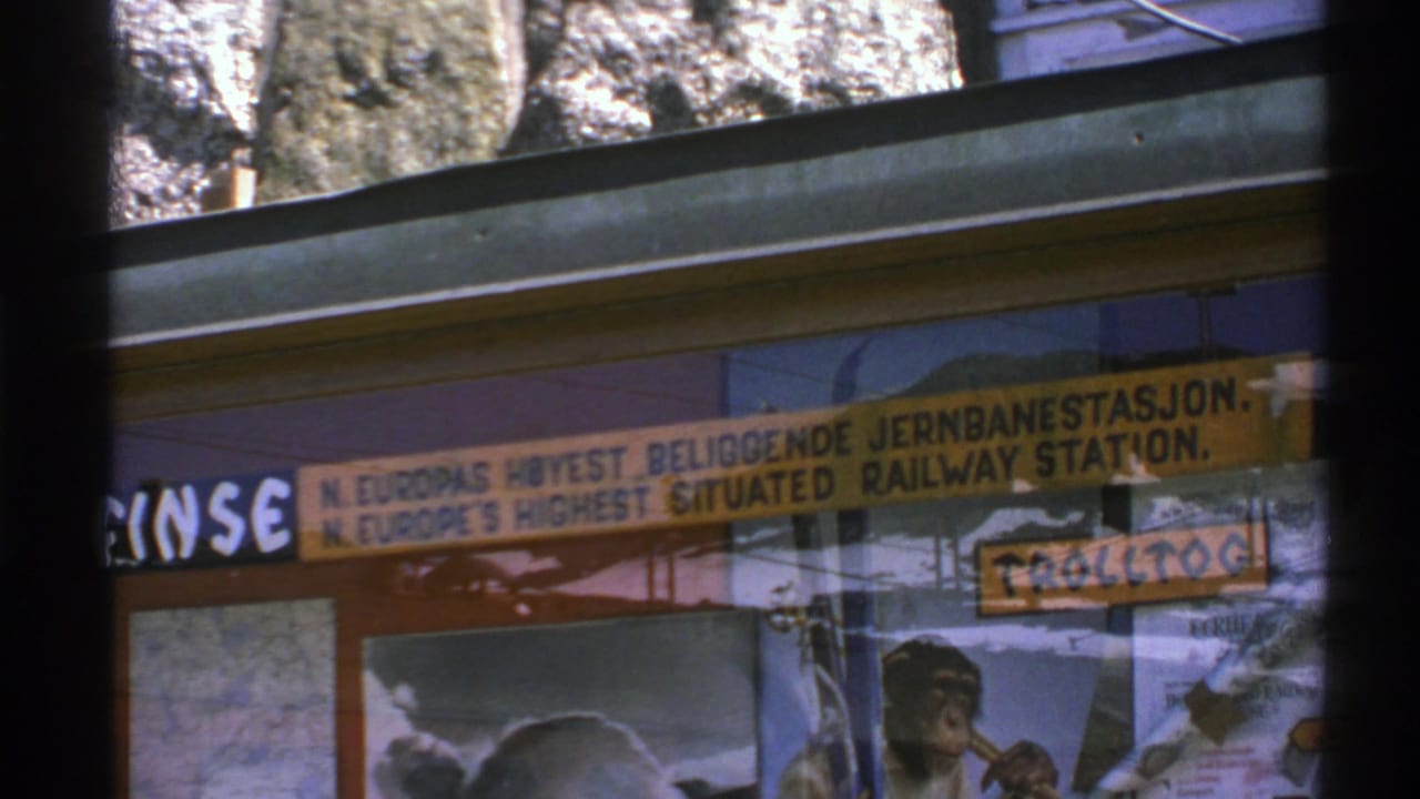1976 Norway home-movie frame showing a high-altitude railway station sign behind dark foreground bars and travel posters.