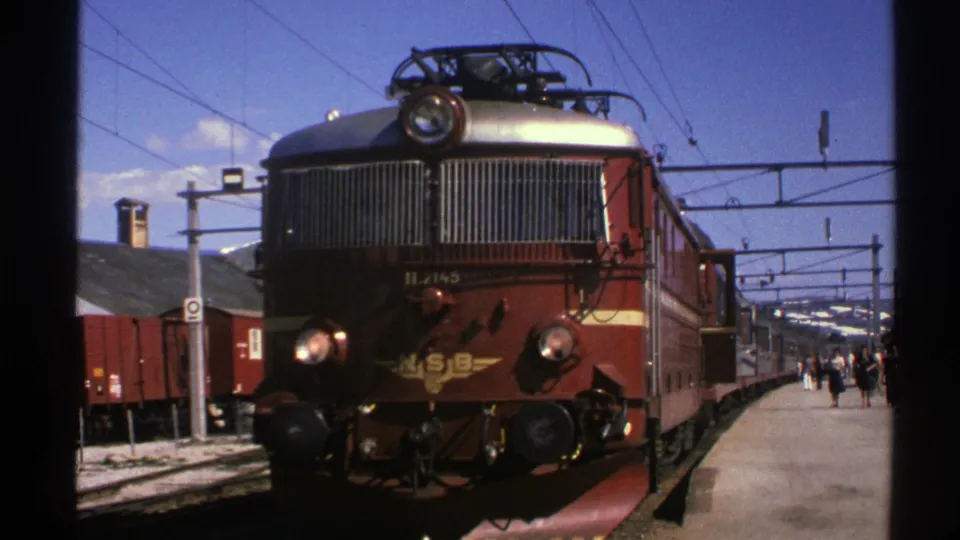 1976 Norway 8mm still with the red NSB electric locomotive closer and slightly darker, framed by wires, railcars, and station platform details.
