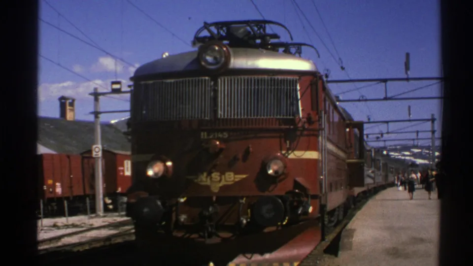 1976 Norway frame showing the stationary red NSB locomotive, platform passengers, electric wires, and railcars in a bright station setting.