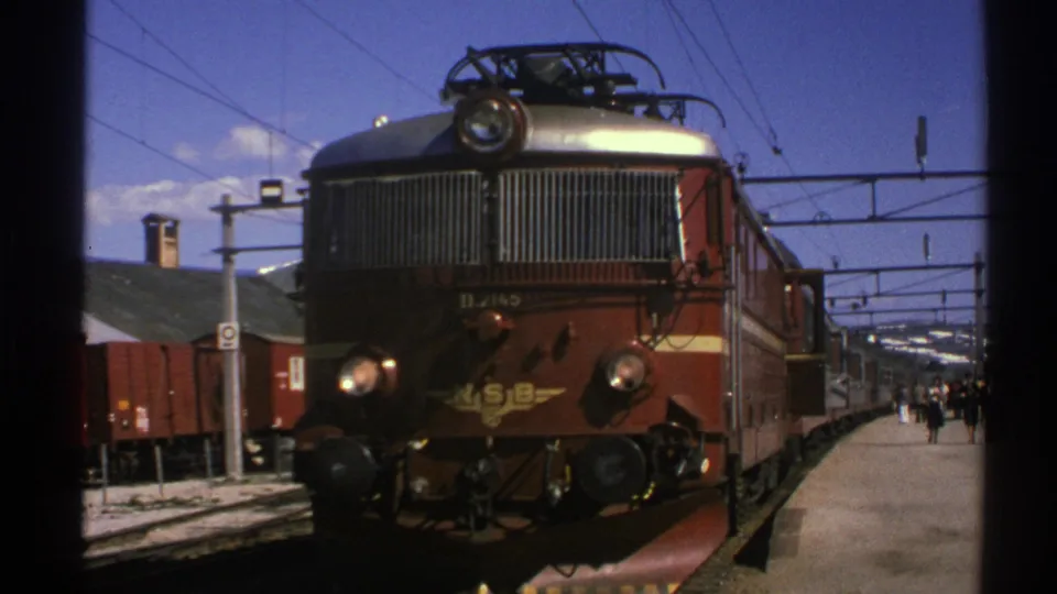 1976 Norway home-movie frame showing a brown and gold NSB electric locomotive at a sunny station platform.