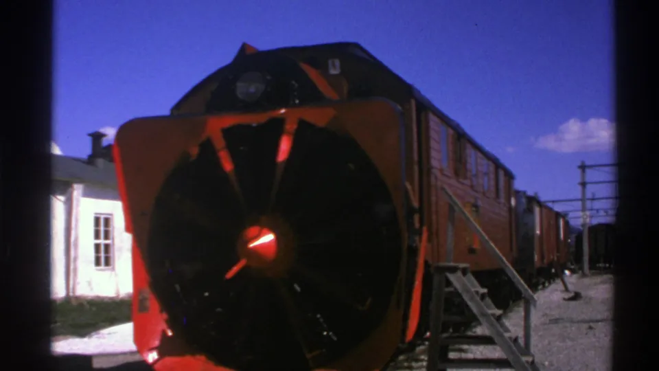 1976 Norway 8mm frame with the red locomotive nose and snowplow dominating the view, with station building and cars behind.