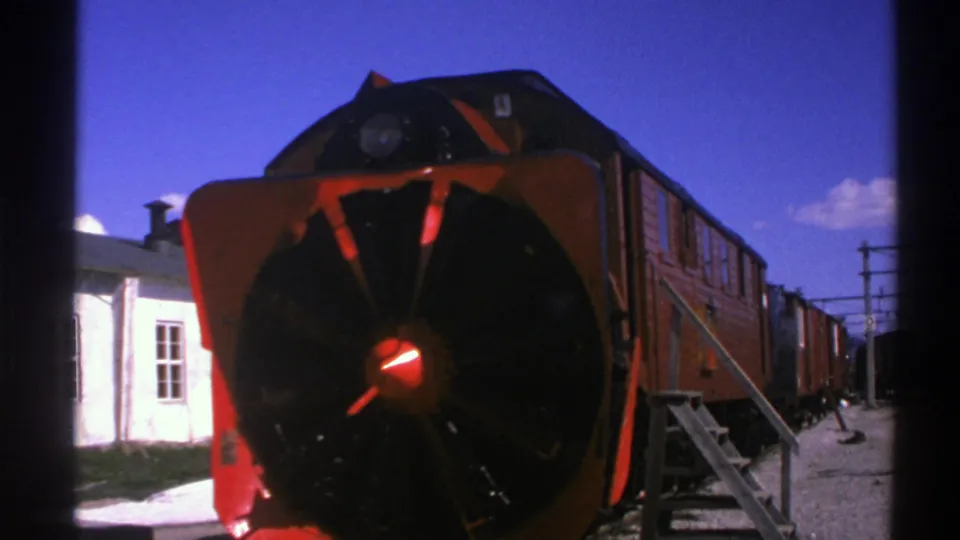 1976 Norway 8mm frame of a red and black snowplow-front train stopped near a white station building and overhead lines.