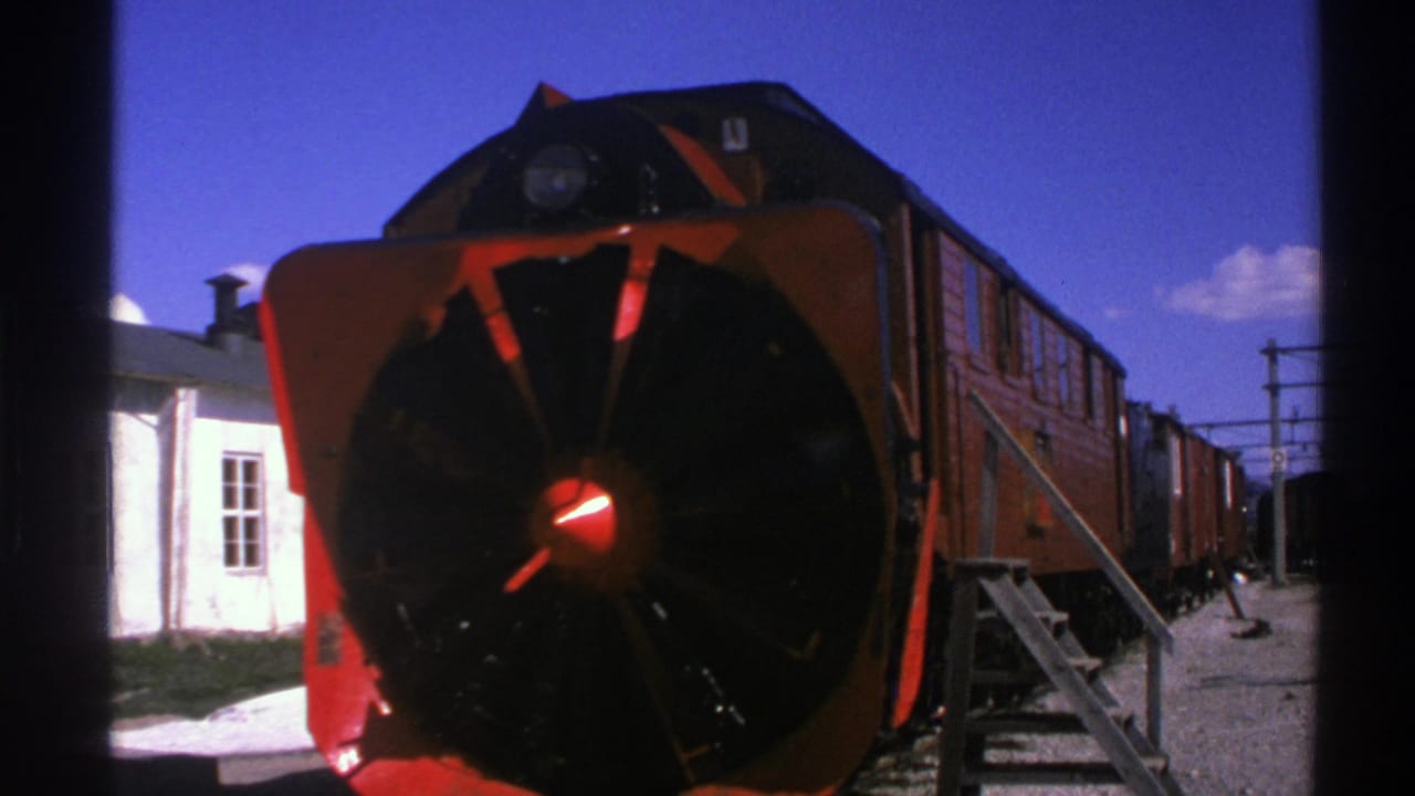 1976 Norway 8mm view of a red locomotive snowplow front, with rail cars receding beside station stairs and a white building.