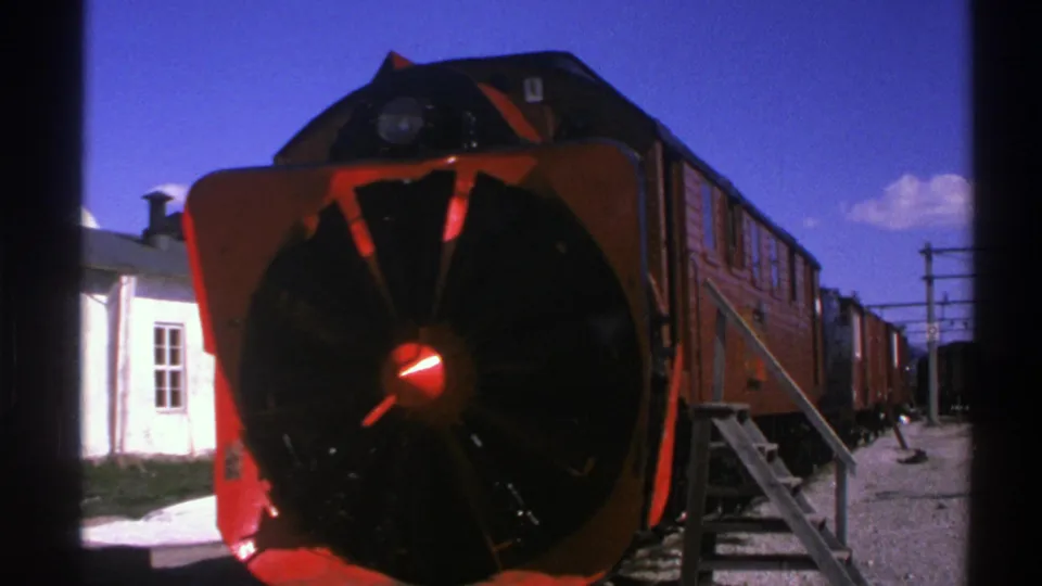1976 Norway 8mm station view with a red snowplow-front train, wooden access stairs, rail cars, and clear blue sky.
