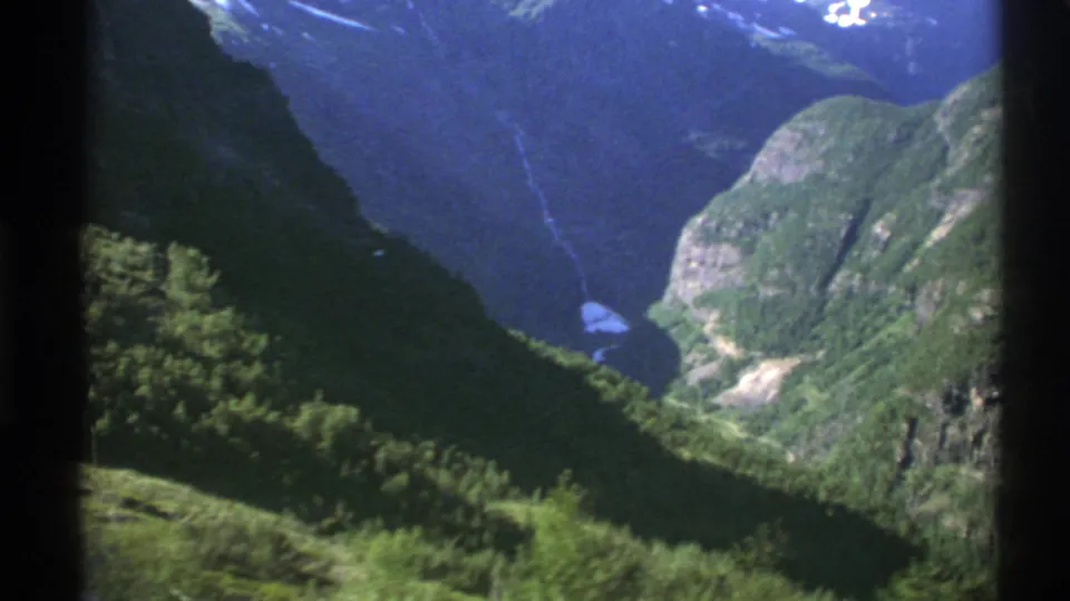 1976 Norway home-movie frame with steep green slopes, a distant fjord or river, and window-edge shadows.
