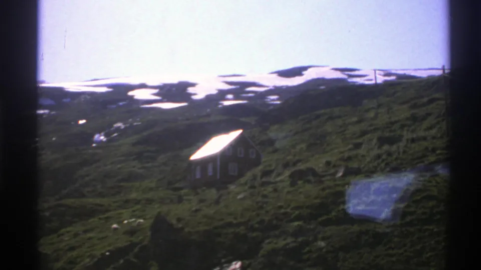 1976 Norway 8mm frame showing a small hillside cabin below snow-streaked alpine terrain.