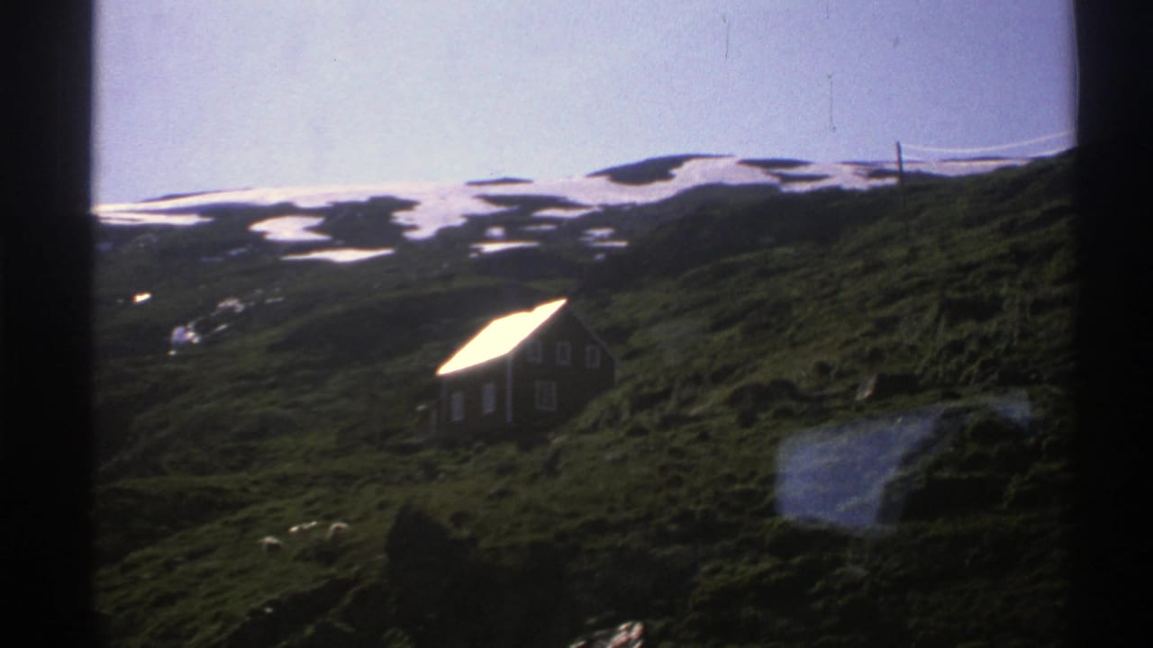 Remote cottage on a green hillside with snow patches beyond.