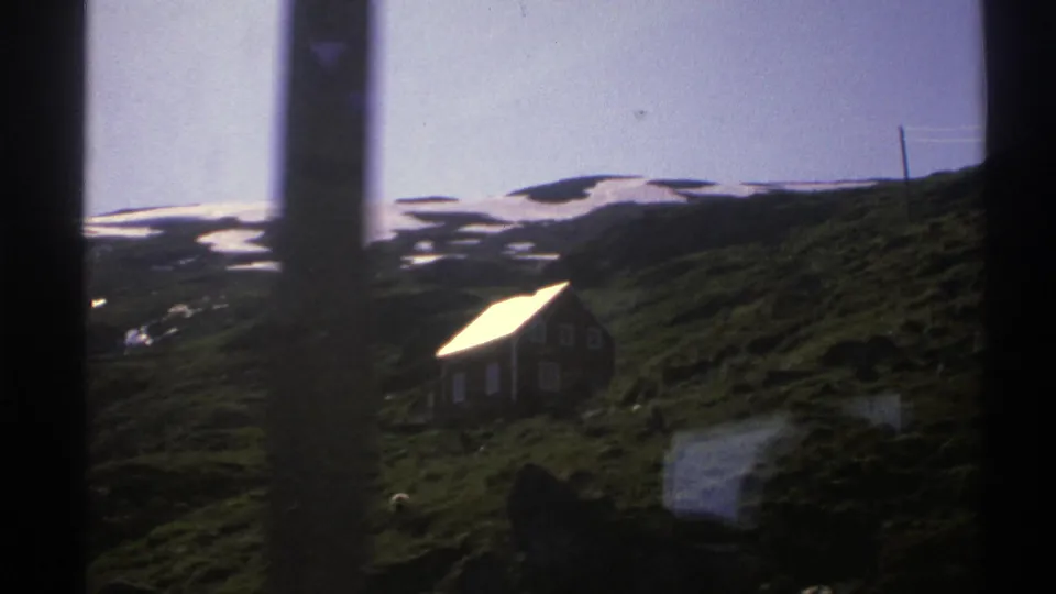 1976 Norway home-movie frame with a hillside cottage, patchy snow in the distance, and vertical film or window-like dark edges.