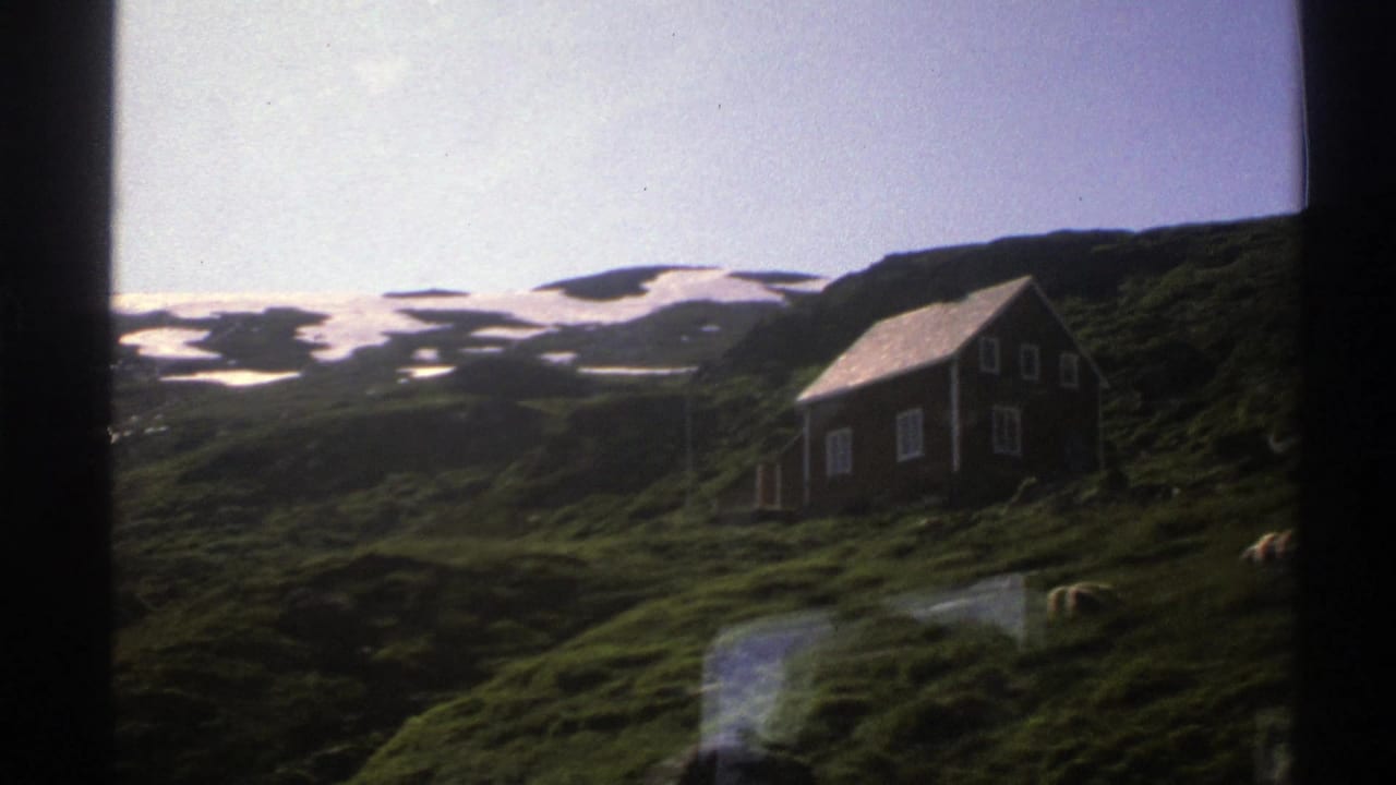 Green Norwegian hillside with a small wooden cabin and patchy snow on distant slopes.