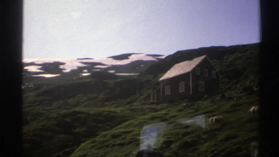 1976 8mm view of a rural Norwegian hillside, showing a small wooden cabin below snow-dappled mountain slopes.