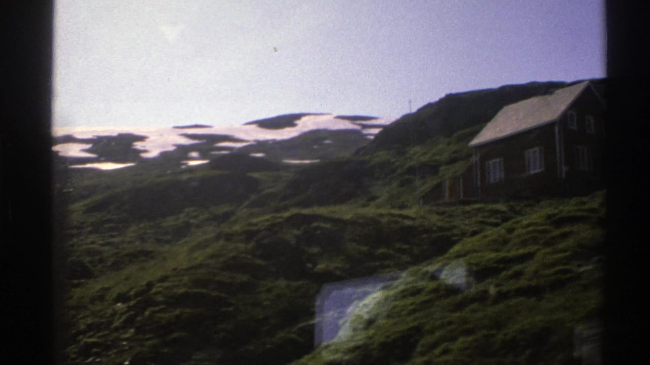Small cabin at the edge of a green slope with snow patches across the hills behind it.