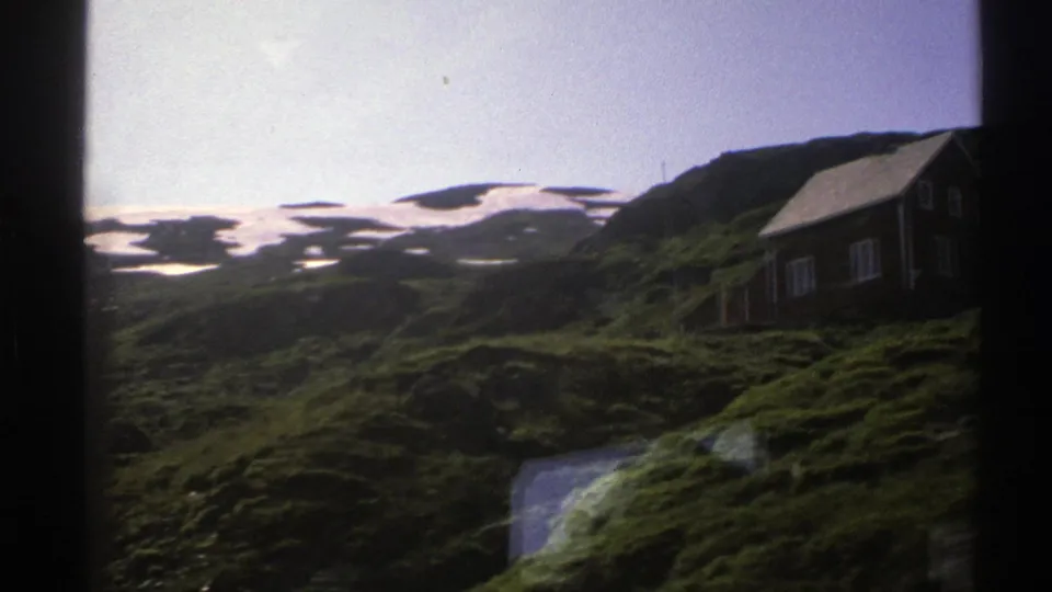 1976 Norwegian home-movie frame showing a wooden cabin on a green hillside with snow patches beyond.