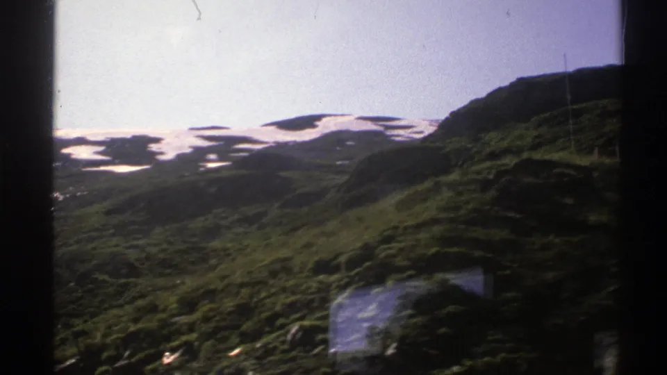 1976 8mm frame of Norwegian mountain terrain, with green foreground slopes and patchy snow in the distance.