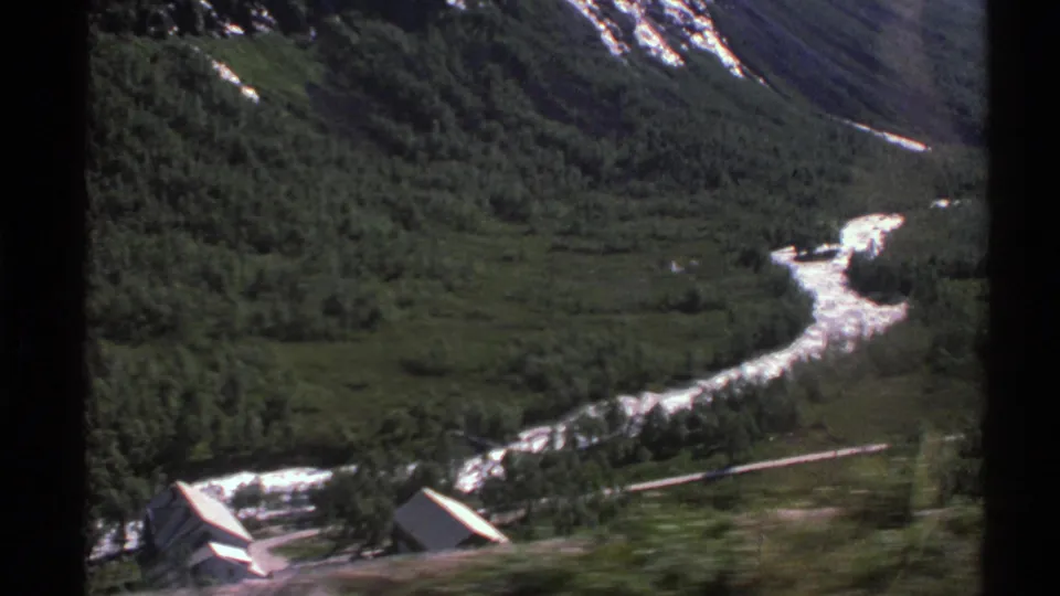1976 Norwegian landscape frame showing a wooded valley, bright winding river, and small rural buildings below steep slopes.