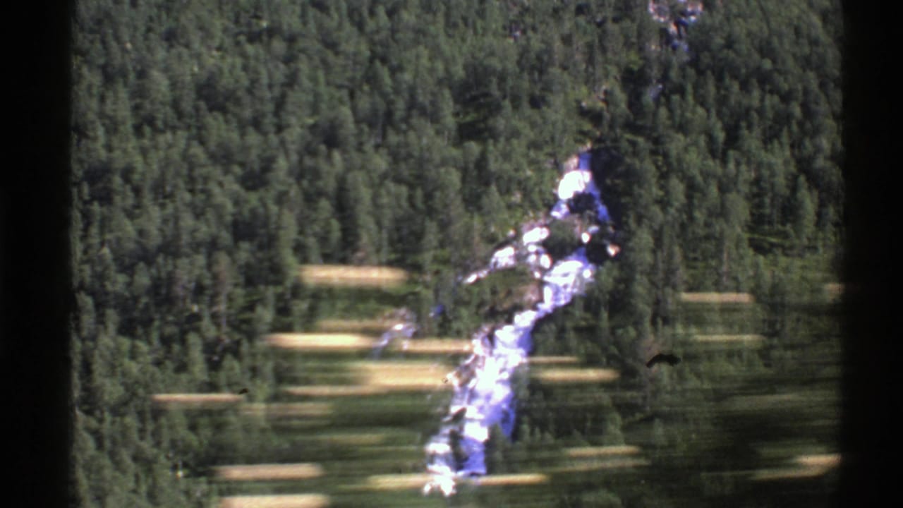 1976 Norway 8mm view of a bright river cutting through dense green forest, framed by dark train-window edges.