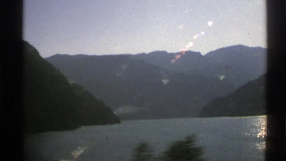 1976 Norway 8mm frame showing a mountain lake, wooded hillsides, distant peaks, and sun glinting on the water from a moving vehicle viewpoint.
