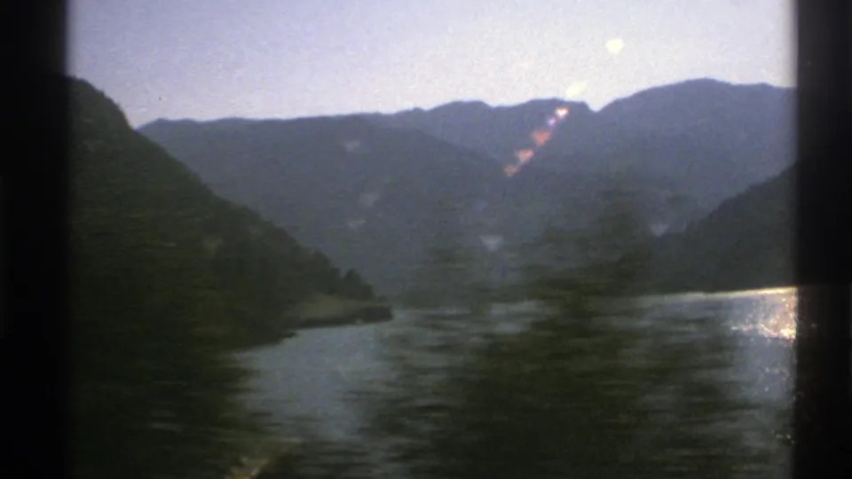 1976 Norway 8mm frame with a lake, dark forested shore, distant mountains, and blurred roadside trees crossing the foreground as the vehicle moves.