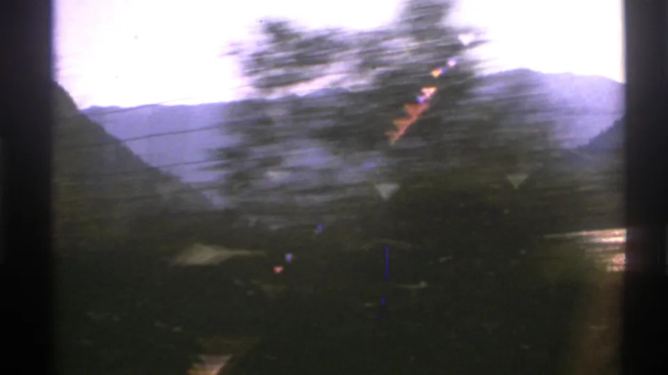 1976 Norway 8mm frame where roadside trees and a possible guardrail blur across a bright lake and mountain backdrop during vehicle movement.