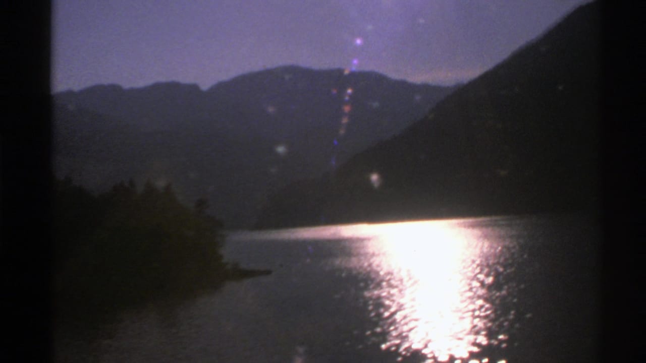 1976 Norway 8mm frame showing a bright sun reflection on a lake, with pine shoreline and silhouetted mountains under a purple sky.