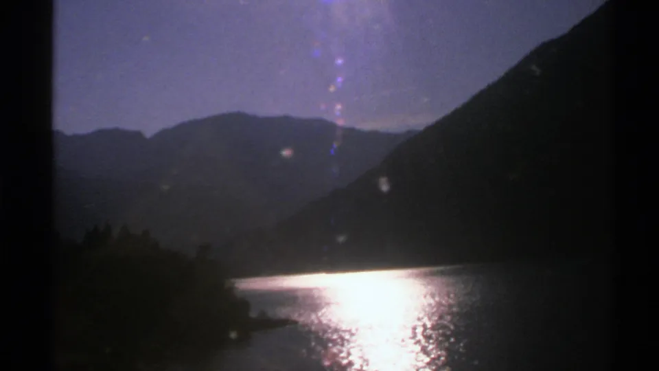 1976 Norway 8mm frame with a lake, strong reflected sunlight, wooded shoreline, and layered mountain silhouettes in dim sky.