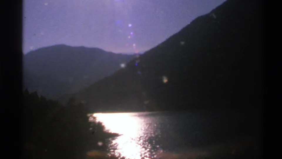 1976 Norway 8mm frame looking over a shaded lake edge toward mountains, with the sun reflection bright on the water.