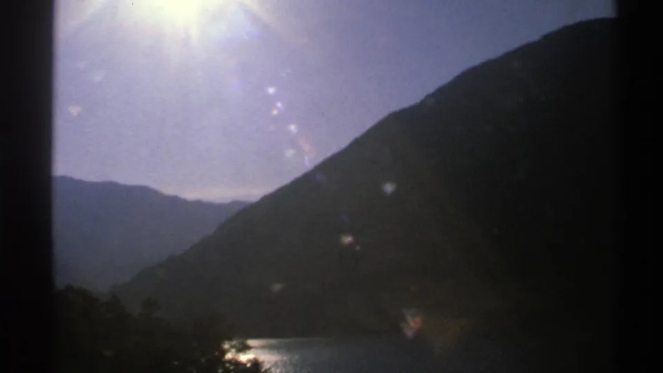1976 Norway 8mm frame with the sun near the top edge, flare across the sky, and a small lake area below dark mountain slopes.