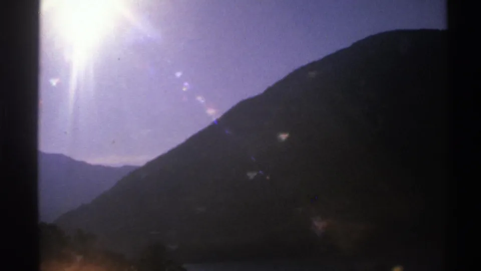 1976 Norway 8mm frame showing a shadowed mountain and pale sky from inside a moving car, with strong sun glare and film grain.