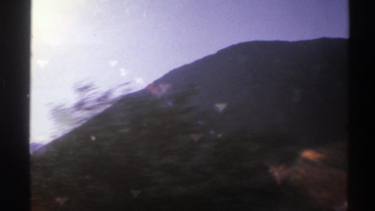 Mountain slope and blurred roadside trees pass outside a car window under a bright sky.