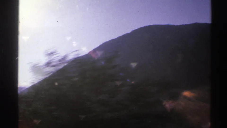 1976 Norway travel frame with a dark mountain ridge, blurred trees in the foreground, and sun-washed sky seen from a moving car.