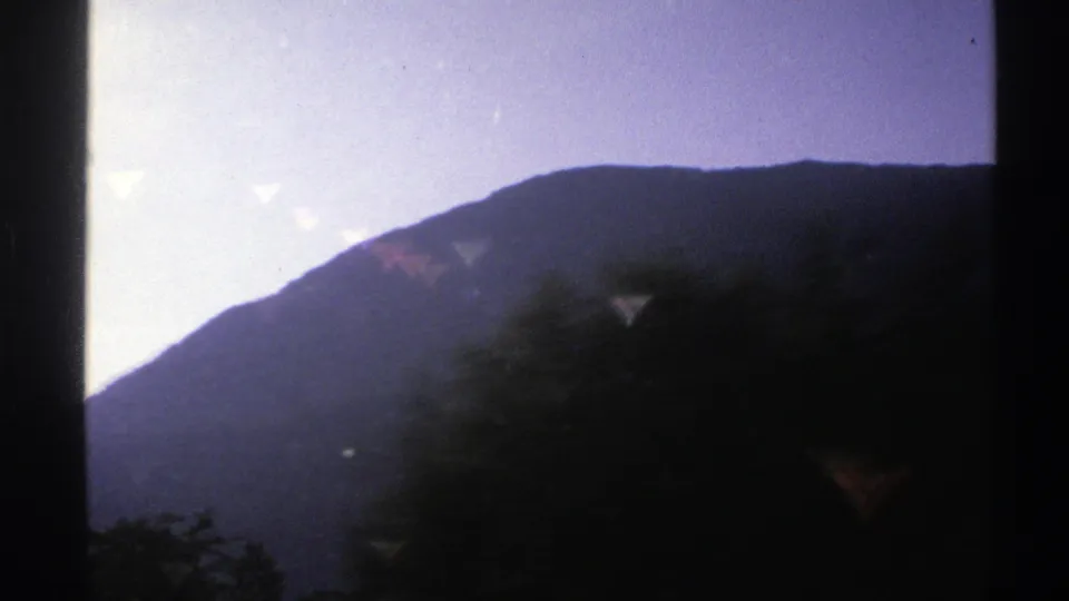 1976 Norway 8mm frame of a broad mountain form seen through a car window, with passing trees blurred across the lower foreground.