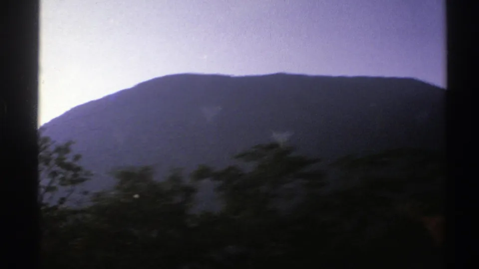 1976 Norway home-movie frame showing a long mountain ridge and roadside vegetation streaking past from inside a moving car.