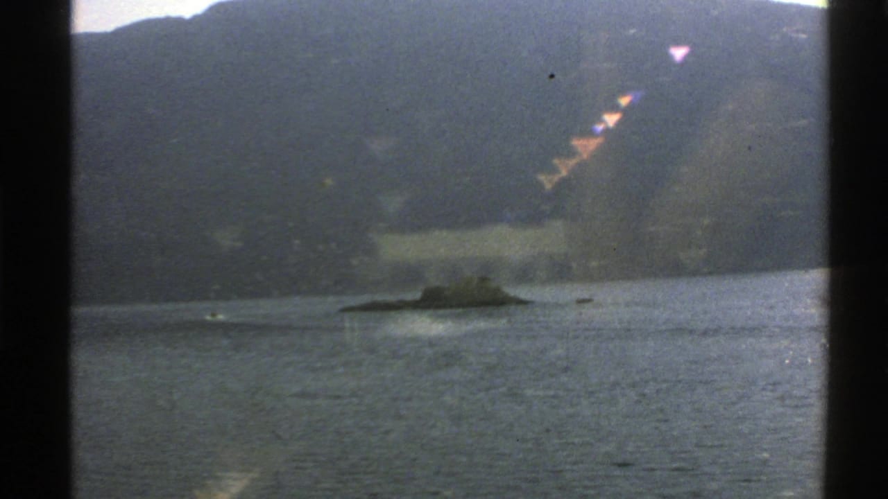 1976 Norway 8mm frame showing a dark submarine-like vessel on calm water, with mountains in the background and a small rocky outcrop nearby.