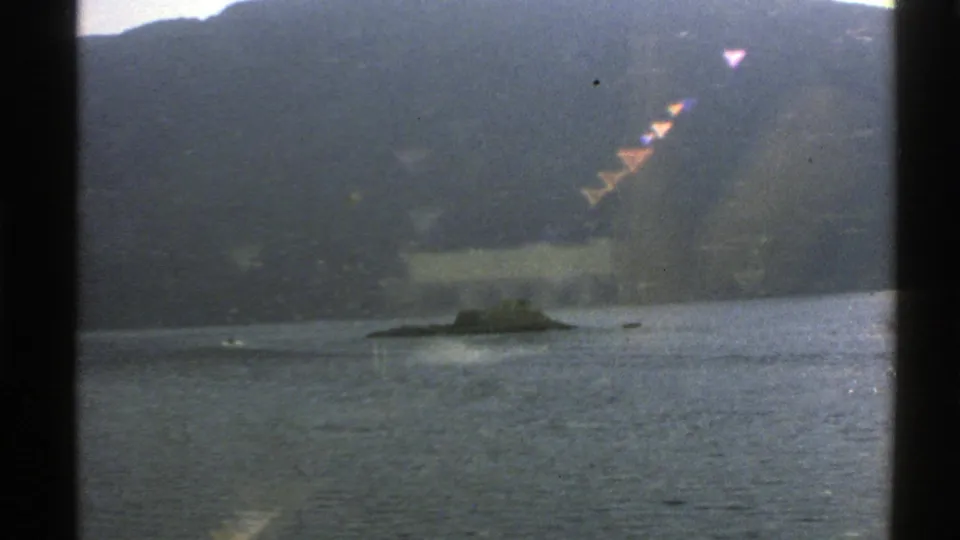 1976 Norway 8mm frame showing a dark submarine-like vessel on calm water, with mountains in the background and a small rocky outcrop nearby.
