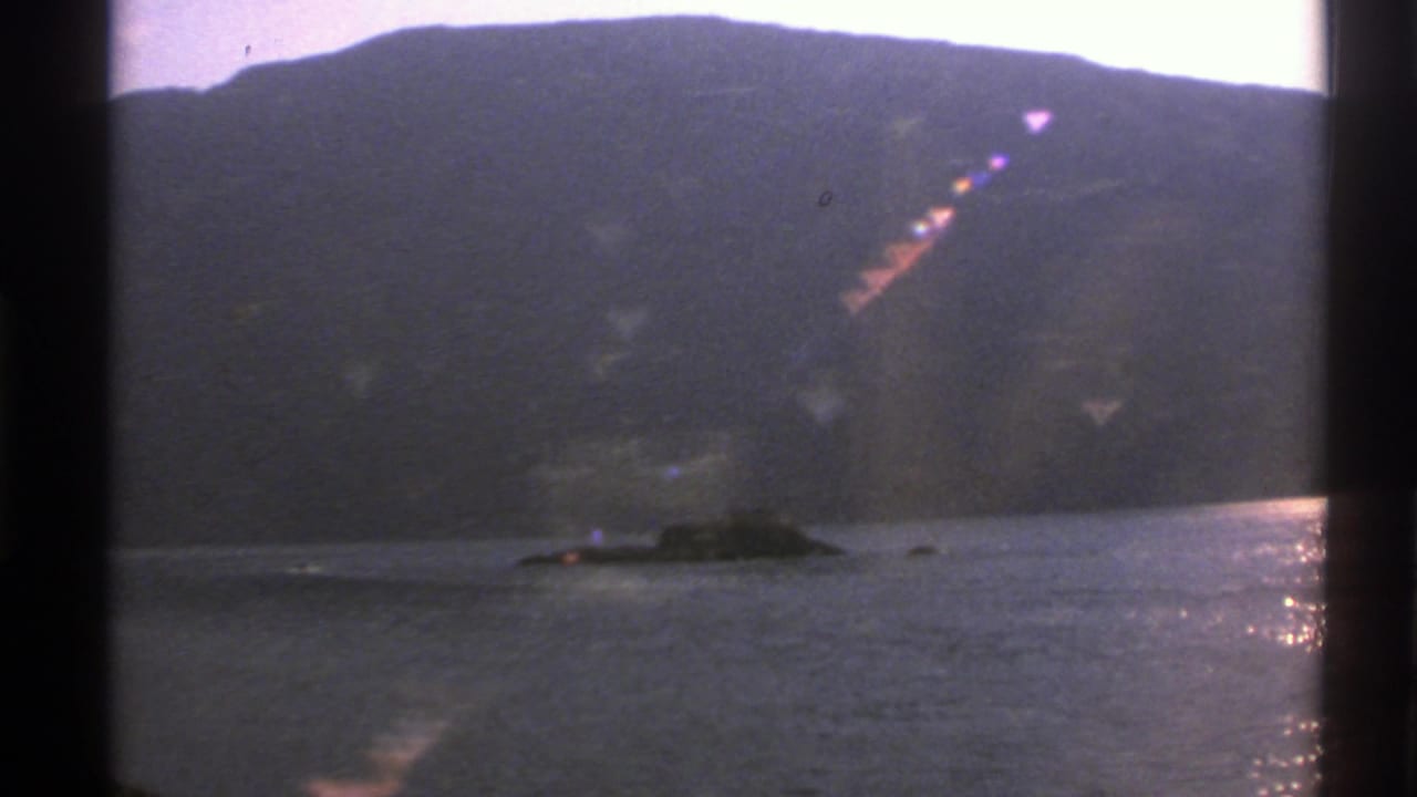 Surfaced vessel near a small rock formation with mountains behind it.