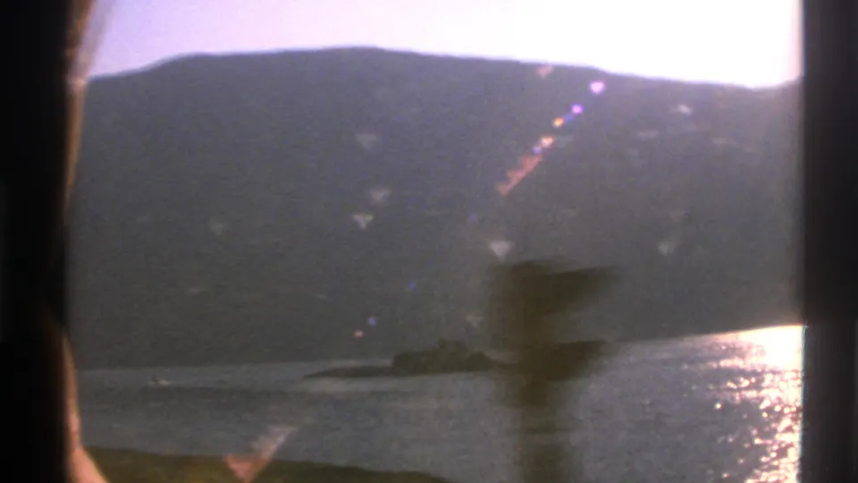 1976 Norwegian coastal 8mm frame with a blurred foreground post, bright water glare, mountains, and a low dark vessel near the shore.
