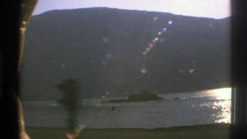Grainy 8mm Norway frame showing a low submarine-like vessel near a rocky outcrop, with mountains, bright water reflection, and film flare.