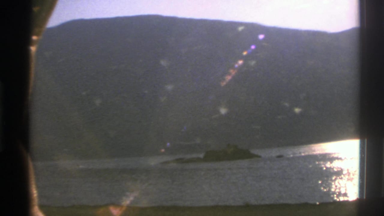 1976 Norway 8mm frame showing a lake, dark mountain ridge, sunlit water reflection, and train window bars.