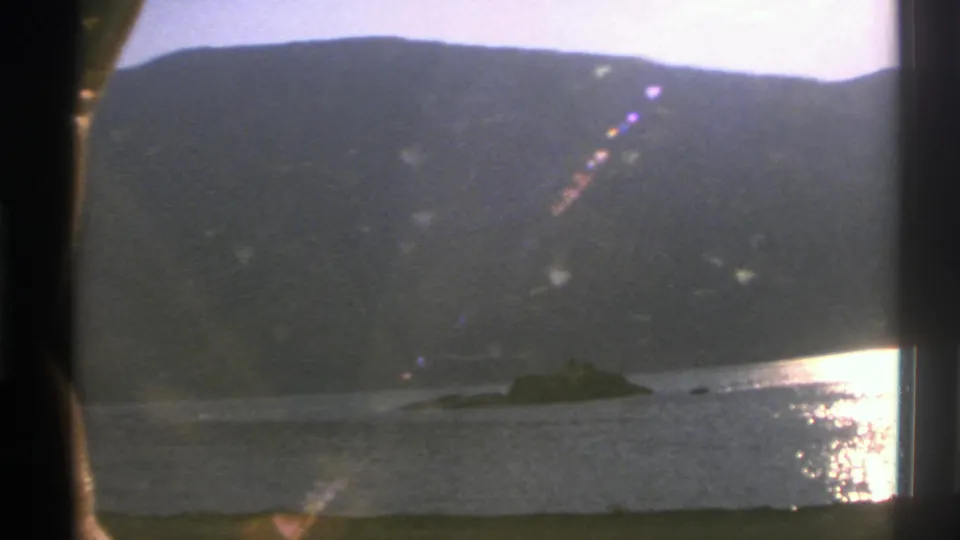 1976 Norway 8mm frame showing a lake, dark mountain ridge, sunlit water reflection, and train window bars.