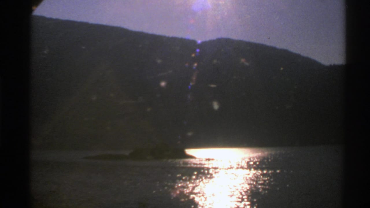 1976 Norway 8mm frame showing a silhouetted mountain ridge, calm lake, and strong sun reflection across the water.