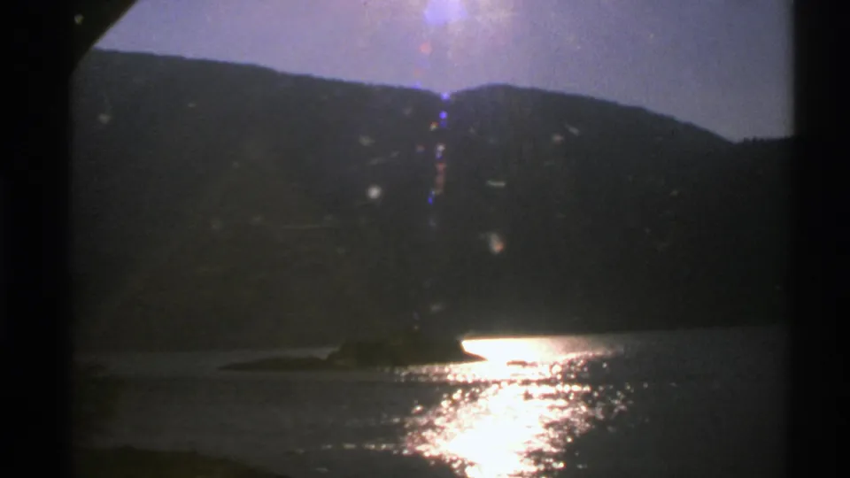 1976 Norway 8mm frame with a dark hillside above a lake, bright reflected sunlight, and visible film-frame borders.