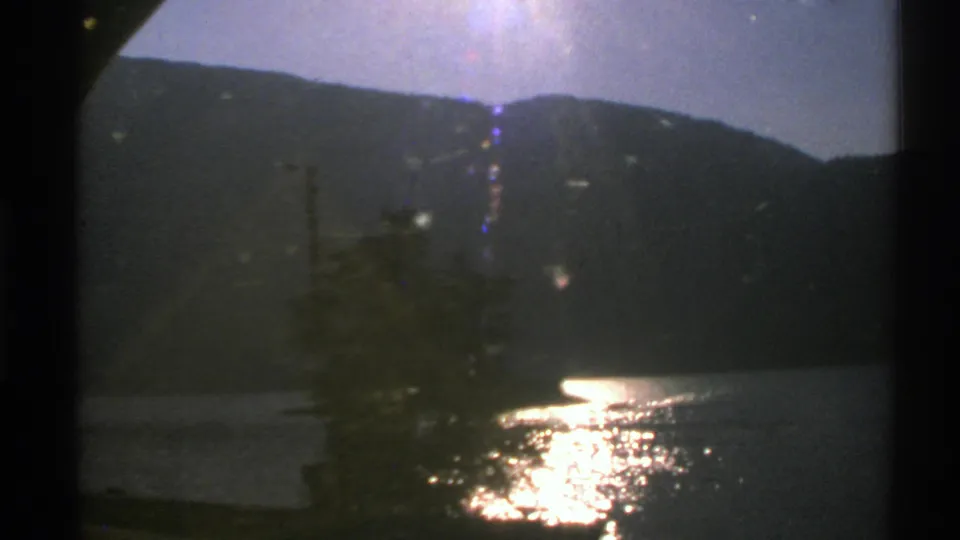 1976 Norway 8mm frame showing foreground trees by a lake, a shadowed mountain ridge, and bright sunlight on the water.