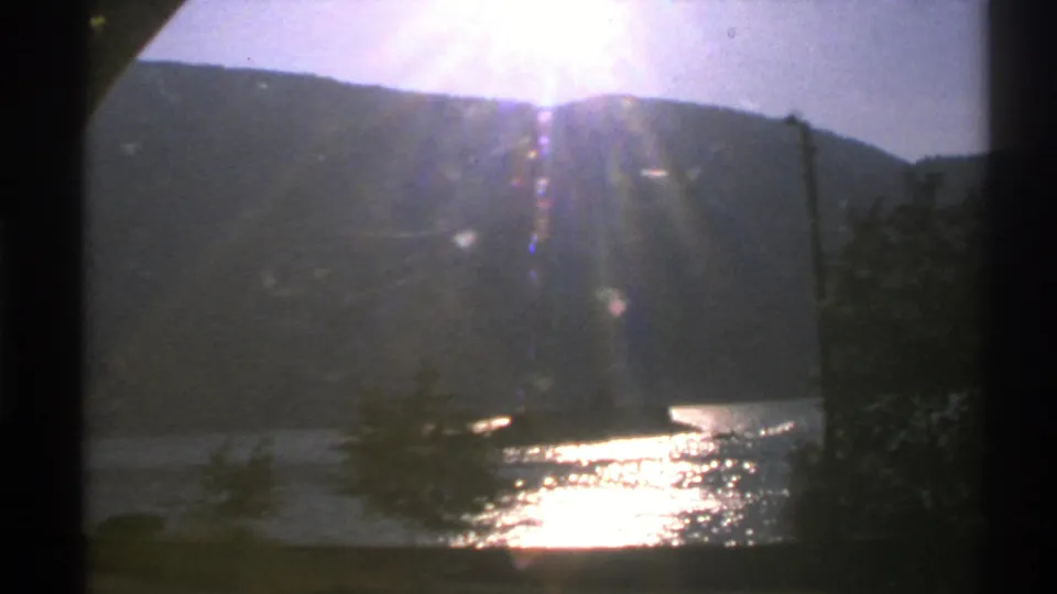 1976 Norway 8mm frame with the sun near the ridge line, lens flare, trees, and a bright reflection across the lake.