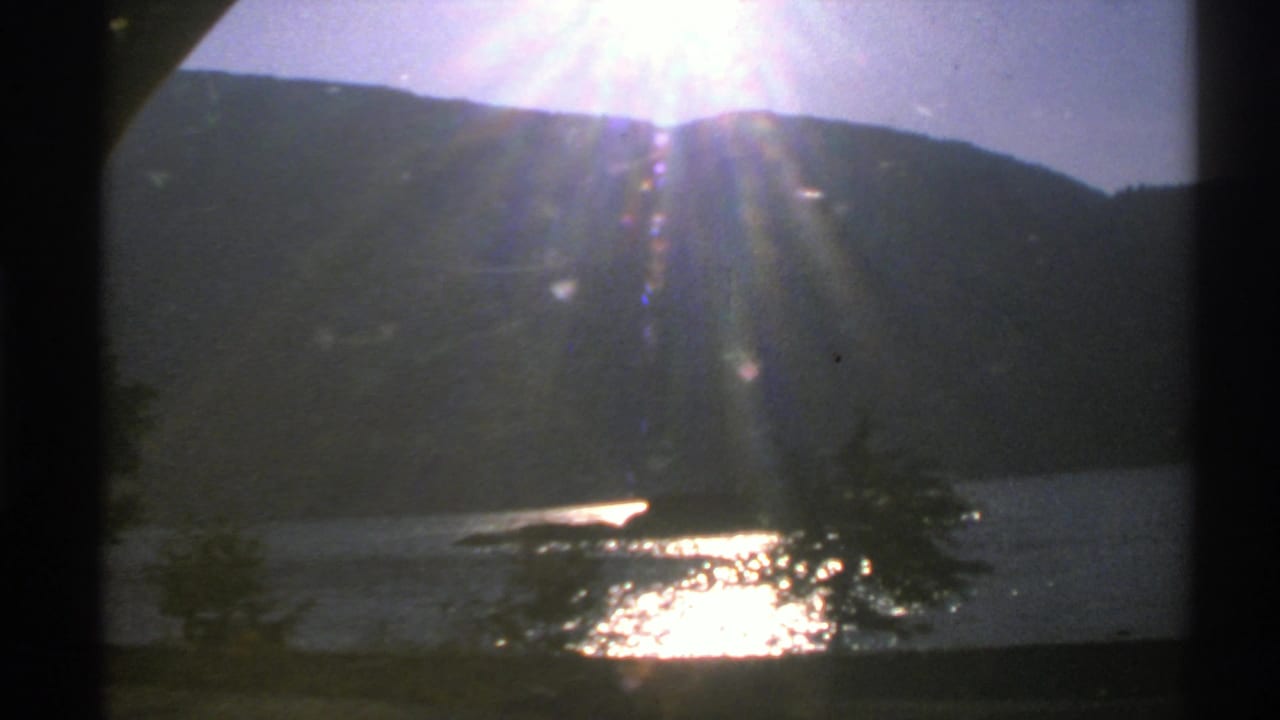 Sun flares over a dark hillside and sparkling lake with trees near the shore.