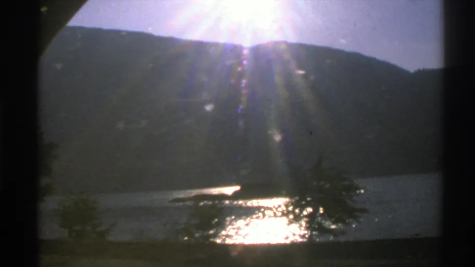 1976 Norway 8mm frame showing bright sun flare above a hillside, reflective lake water, shoreline trees, and dark film-frame edges.