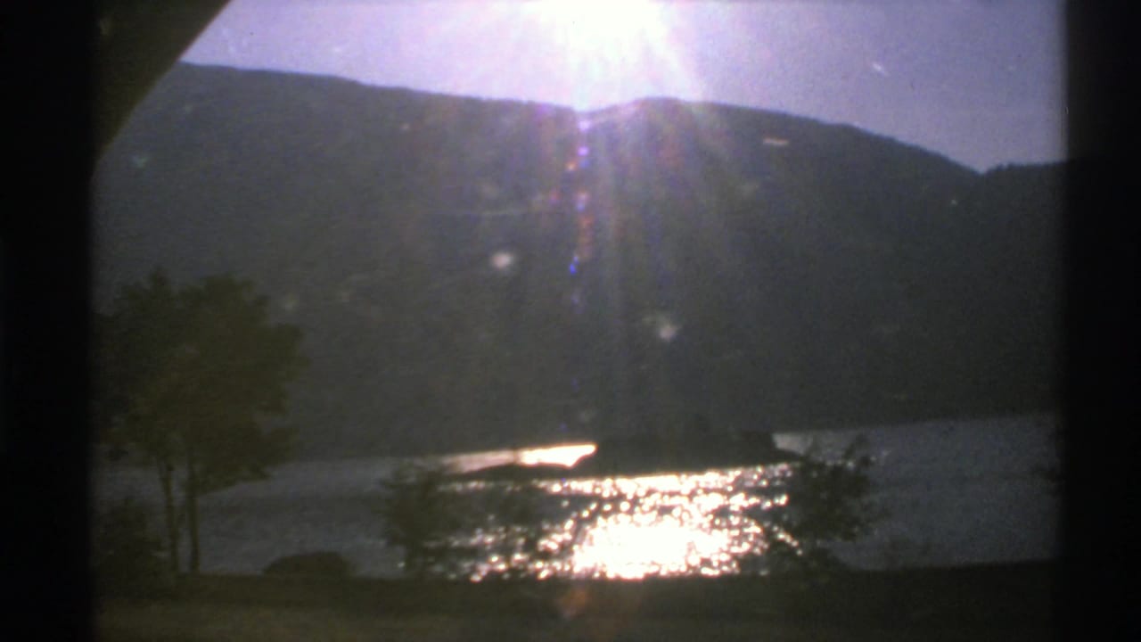1976 Norway 8mm frame with a dark mountain slope, bright reflection on the lake, small shoreline trees, and a narrow landform in the water.