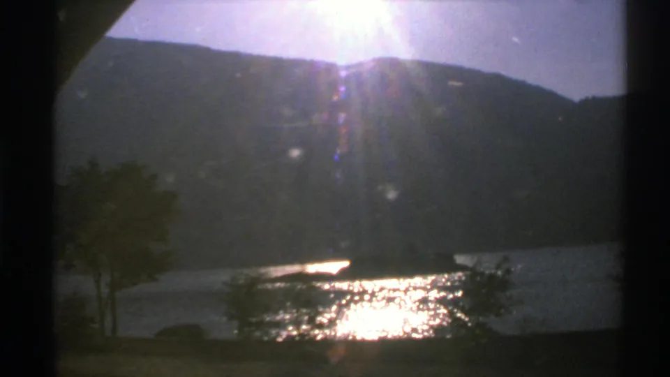 1976 Norway 8mm frame with a dark mountain slope, bright reflection on the lake, small shoreline trees, and a narrow landform in the water.