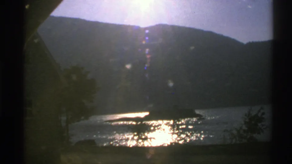 1976 Norway 8mm frame showing shimmering water, a dark hillside, near-shore trees, foreground vegetation, and strong flare from the sun.